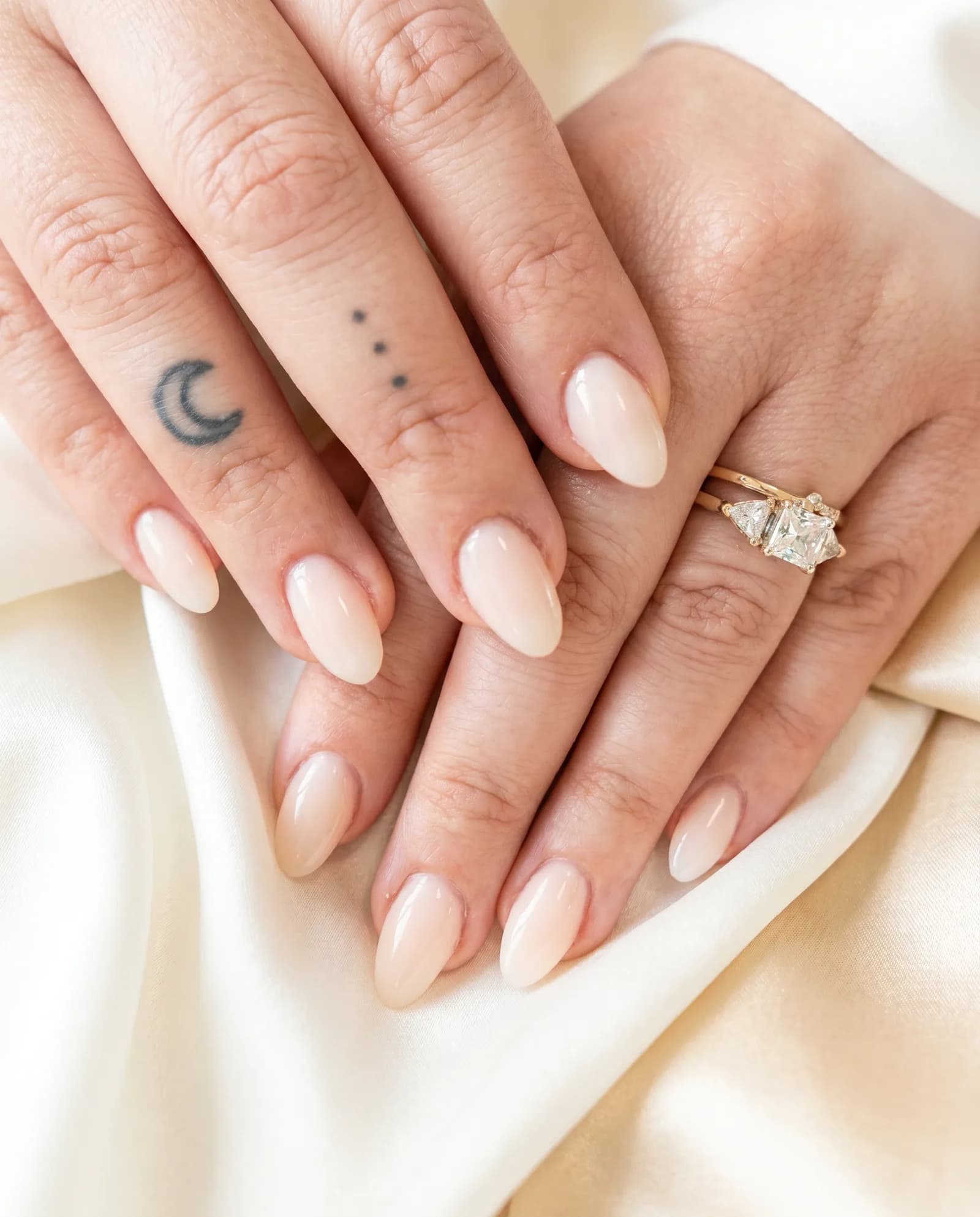 Nude almond manicure and a diamond engagement ring resting on soft ivory-champagne silk.